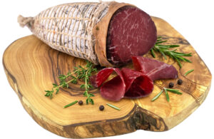 Whole cured ham leg on a wooden cutting board, tied with nets, with rosemary sprigs and peppercorns nearby and several slices showing.