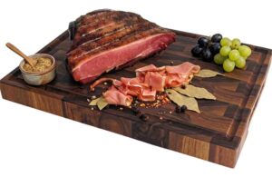 Large roasted ham on a wooden cutting board with sliced ham, grapes, bay leaves, peppercorns and a small bowl of mustard.